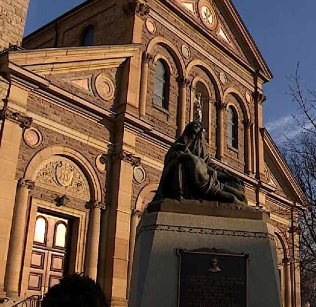 Sculpture d’une femme qui tient un homme mourant dans ses bras. Statue installée sur un socle de couleur grise. La façade d’une église est aperçue à l’arrière-plan.