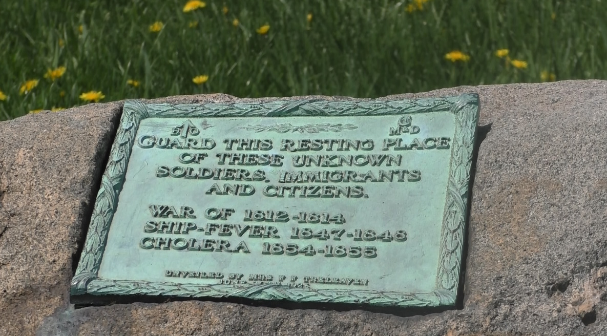 Inscription sur une plaque verte et rectangulaire, installée sur un imposant socle de pierre de couleur grise. De l’herbe verte est aperçue à l’arrière-plan.