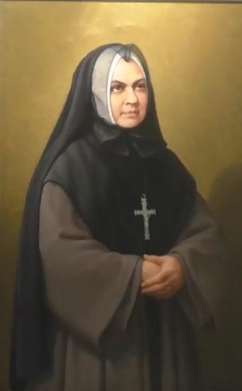Colour portrait of middle aged woman wearing a veil, dark nun’s habit and a large silver cross, looking away from viewer. 