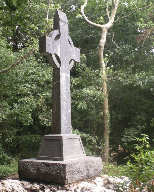 Photographie d’une croix celtique de couleur grise, installée sur un socle et vue de biais. Des arbres sont visibles à l’arrière-plan.