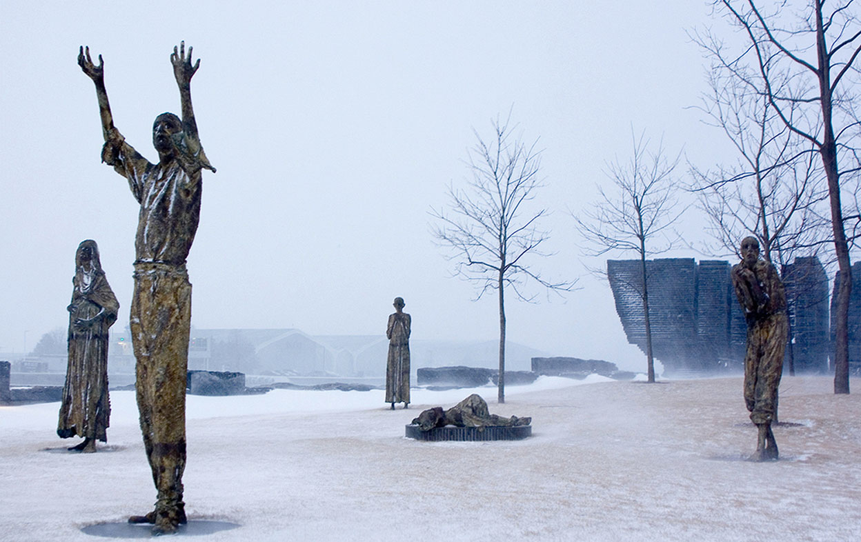 Quatre statues se tenant debout, dont celle d’un homme levant les bras en l’air, ainsi qu’une cinquième statue, couchée par terre. Le mur de pierre sculptées est visible à l’arrière-plan. Le sol est couvert de neige et trois arbres sans feuilles sont visibles derrière les sculptures.