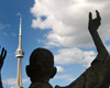 Rear view of upper torso and back of head of male sculpture with arms upraised, facing CN Tower in background. Blue sky with clouds.