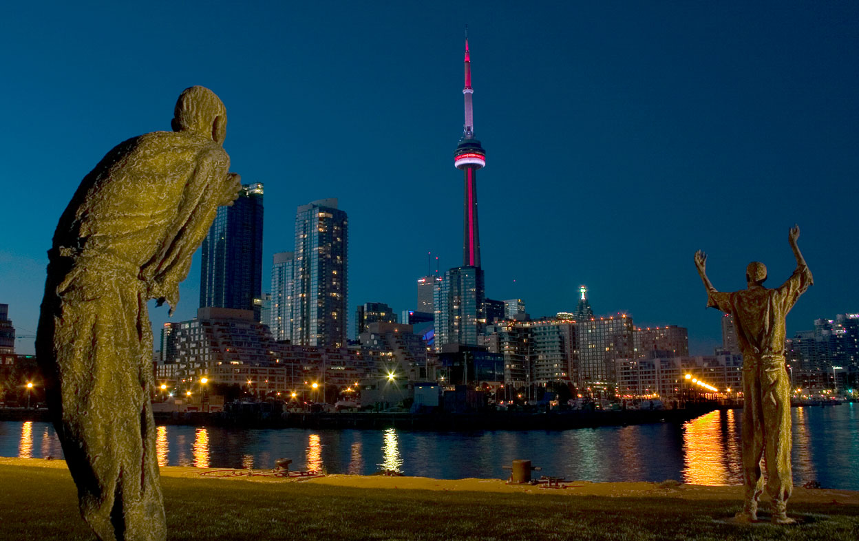 Night scene of two sculptural figures viewed from behind, with one on left hunched over and one on right with arms upraised facing CN Tower.  City skyline harbour in background.