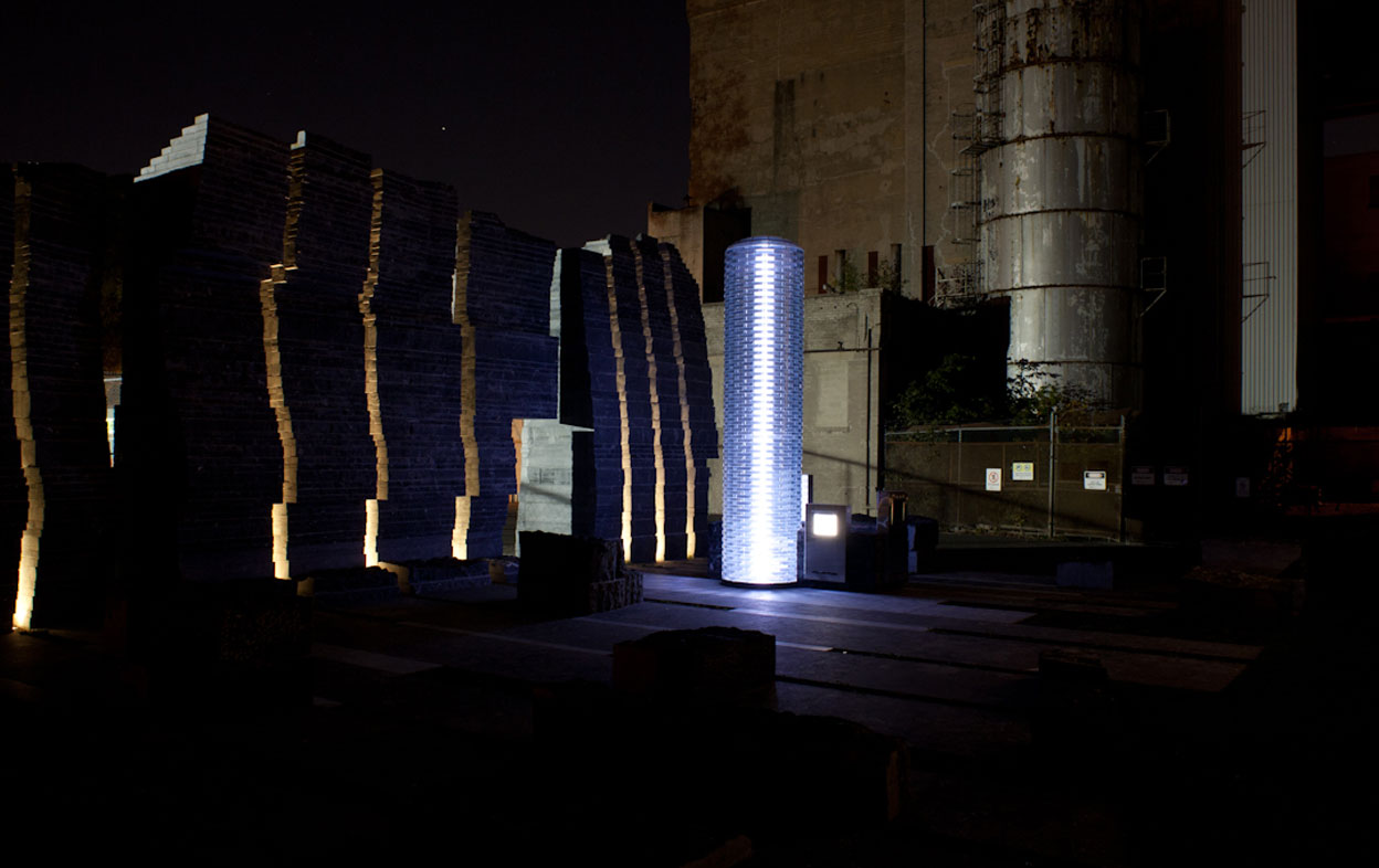 Night scene of lit up glass cylinder beacon, with illuminated columns of stone wall on left, large cement building in right background.