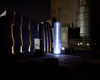 Night scene of lit up glass cylinder beacon, with illuminated columns of stone wall on left, large cement building in right background.