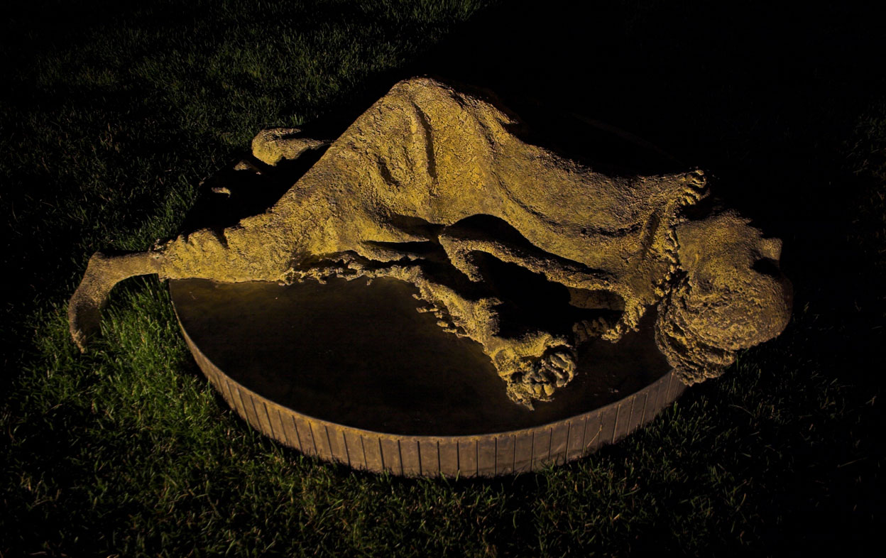 Night scene of sculptural figure of woman lying down on the ground, propping herself up on her arms and legs.  Sculpture mounted on circular plinth.
