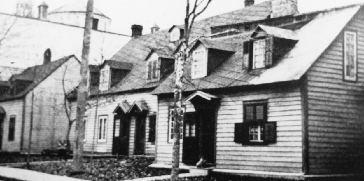 Une photographie en noir et blanc de deux bâtisses en bois aperçues de la rue. Les toits en pente, les fenêtres à pignon, une église et deux arbres sont bien visibles.