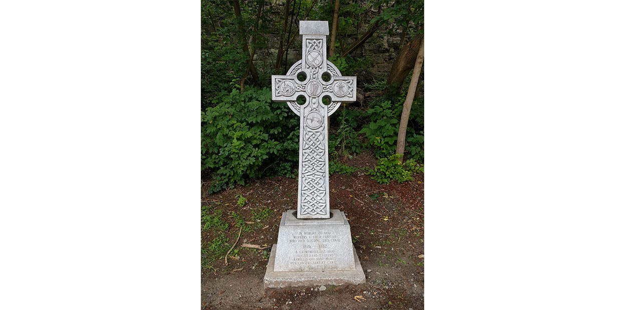 Croix celtique de couleur grise, vue de face. Des ornements façonnent sa structure, de la terre et du feuillage vert sont aperçus à l’arrière-plan.