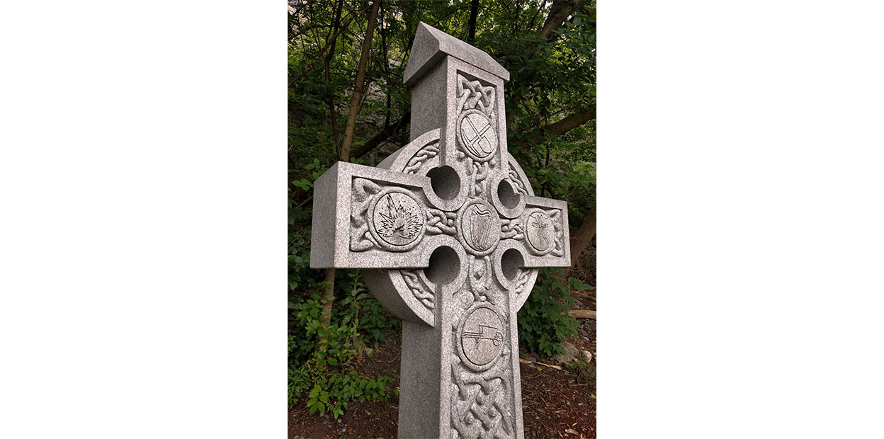 Slightly diagonal view of top of  grey Celtic Cross with icons clockwise from of pick and shovel, mosquito, wheelbarrow, and explosion.
