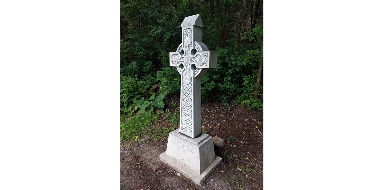 Vue diagonale du monument de la croix celtique aux Irlandais du Canal Rideau. Feuillage verdoyant à l’arrière-plan.