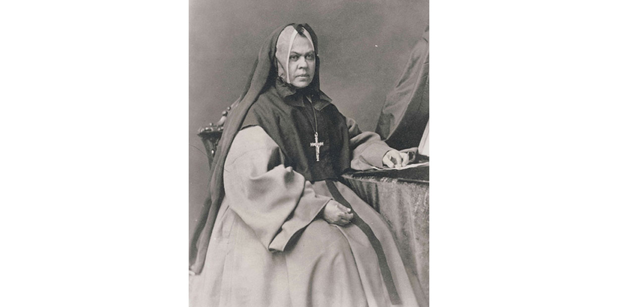 Black and white portrait of seated middle aged woman wearing a nun’s habit and a large silver cross with full skirt, viewed diagonally.