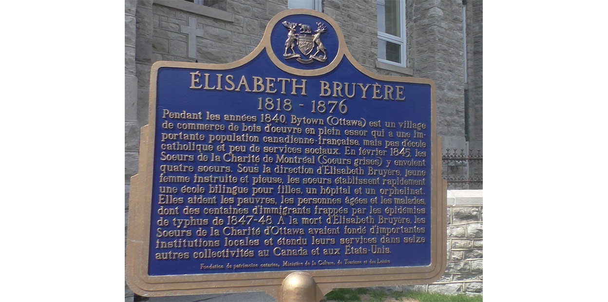 Blue historical plaque titled Élizabeth Bruyère. 1818-1876, with a paragraph written in French about her below. Grey brick wall and building, windows, visible in background.