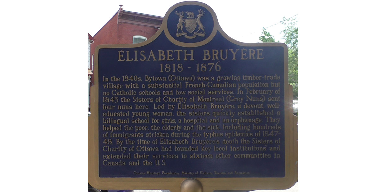 Blue historical plaque titled Élizabeth Bruyère. 1818-1876, with a paragraph written in English about her below. It reads: In the 1840s, Bytown  (Ottawa) was a growing timber trade village with a substantial French-Canadian population but no Catholic schools and few social services. In February of 1845 the Sisters of Charity of Montreal (Grey Nuns) sent four nuns here. Led by Élizabeth Bruyère, a devout, well educated young woman, the sisters quickly established a bilingual school for girls, a hospital and an orphanage. They helped the poor, the elderly and the sick, including hundreds of immigrants stricken during the typhus epidemic of 1847-48. By the time of Élizabeth Bruyère’s death the Sisters of Charity of Ottawa had founded key local institutions and extended their services to sixteen other communities in Canada and the U.S. Street and house visible in background.