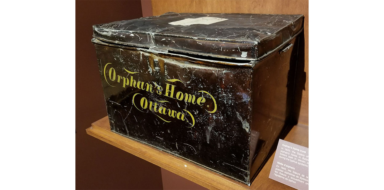 Diagonal view of large, scuffed dark metal chest with Orphan’s Home and Ottawa written on it in yellow script. It is displayed on a brown wooden shelf with grey interpretive note beside it.