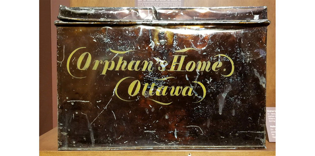 Vue avant d’un grand coffre de métal foncé égratigné où il est écrit Orphan’s Home et Ottawa, en jaune.