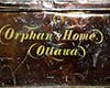 Frontal view of large, scuffed dark metal chest with Orphan’s Home and Ottawa written on it in yellow script.