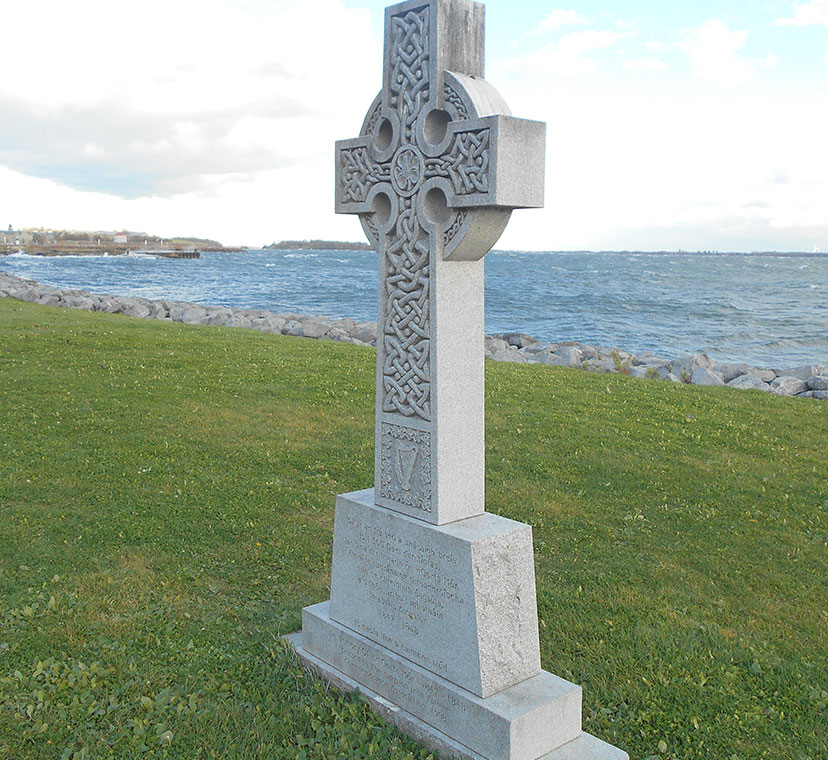 Diagonal View of Celtic Cross on green grass at waterfront, expansive lake in background.
