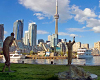 Wide angle image with one sculptural figure lying down in foreground, three standing sculptural figures in middle ground, and city skyline and CN Tower in background across an expanse of water, under a blue sky with white clouds.