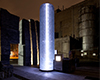 Beacon of light single column of glass lit up at night, surrounded by sculptural stone wall in background. Stone floor stone slab tiles in foreground.