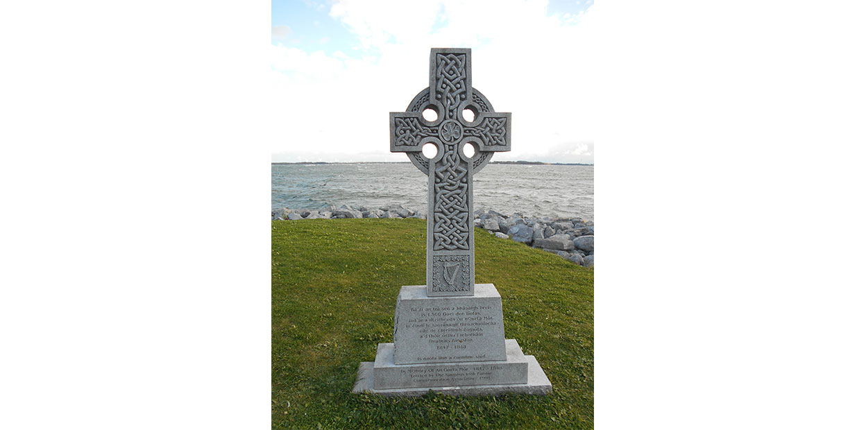 Frontal view of grey Celtic Cross with ornate patterning and inscription on plinth, on green grass with stones on shore, choppy water, and blue cloudy sky visible in background.