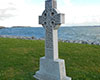 Diagonal view of grey Celtic Cross with ornate patterning and inscription on plinth, on green grass with stones on shore, choppy water, green land masses and blue cloudy sky visible in background.