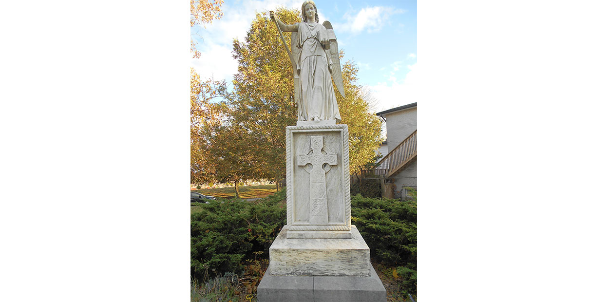 Photographie d’une sculpture de couleur blanche, représentant une statue angélique qui tient un livre ouvert en ses mains. La statue fait face au visiteur.