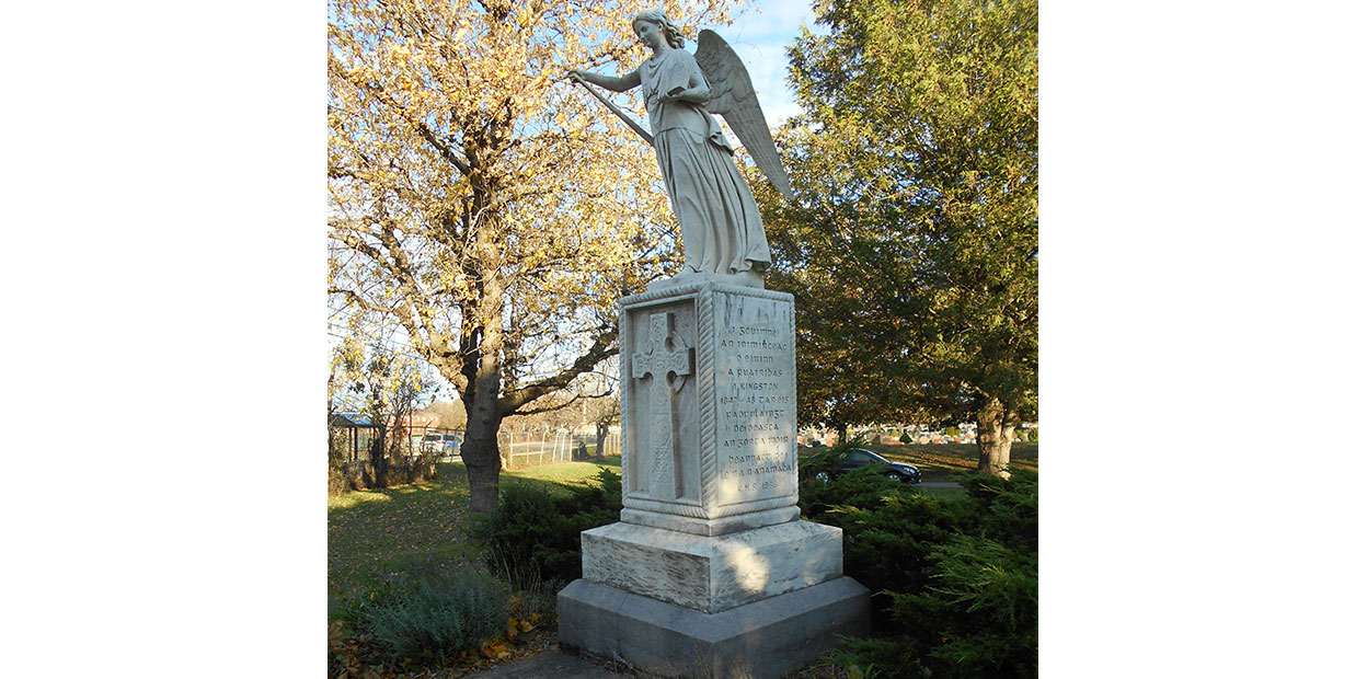 Frontal view of white sculpture of an angelic woman holding an open book standing on plinth and facing viewer. She is standing on a large plinth with an engraved cross, with green bush, tree, and part of a house visible in background.