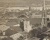 Black and white photo of street from above, first three storey building on left in three quarters profile, adjacent to small church with square tower, four houses, and much larger church with soaring steeple, city skyline and lake in background.
