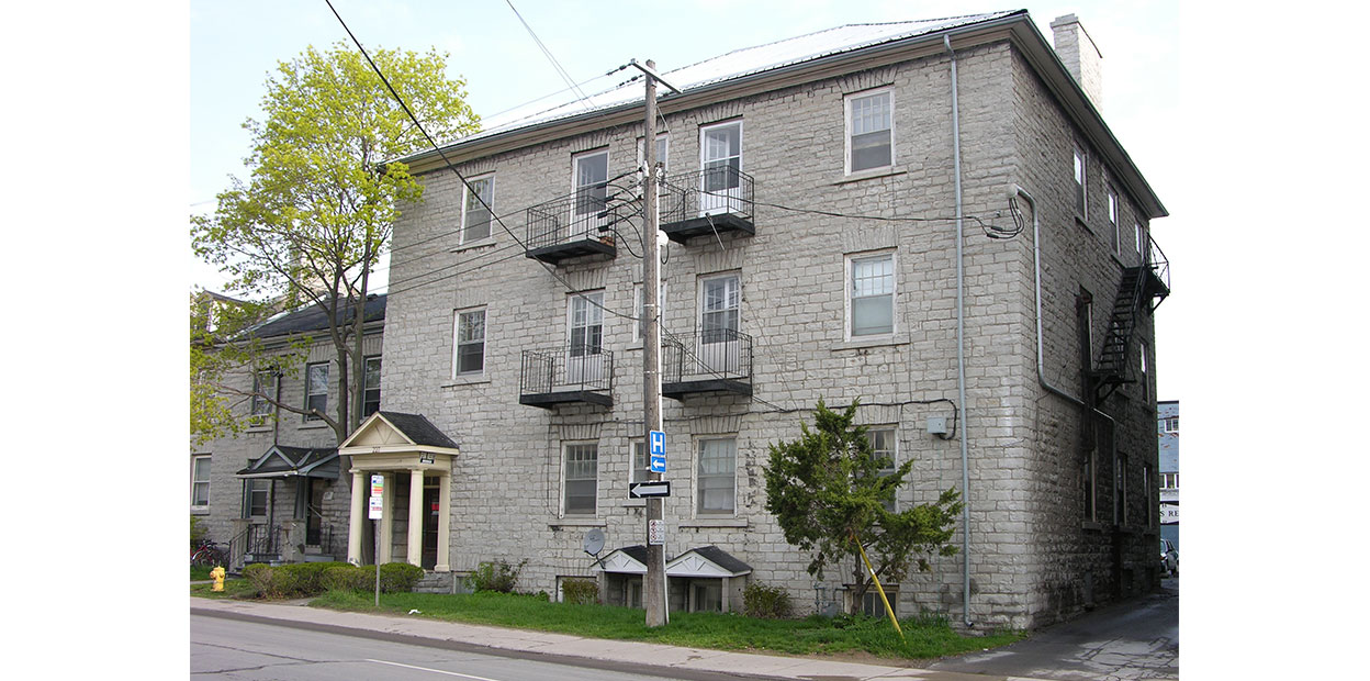 Colour photo, front profile of three storey grey stone building, four windows and two balconies on upper floors, viewed from street.