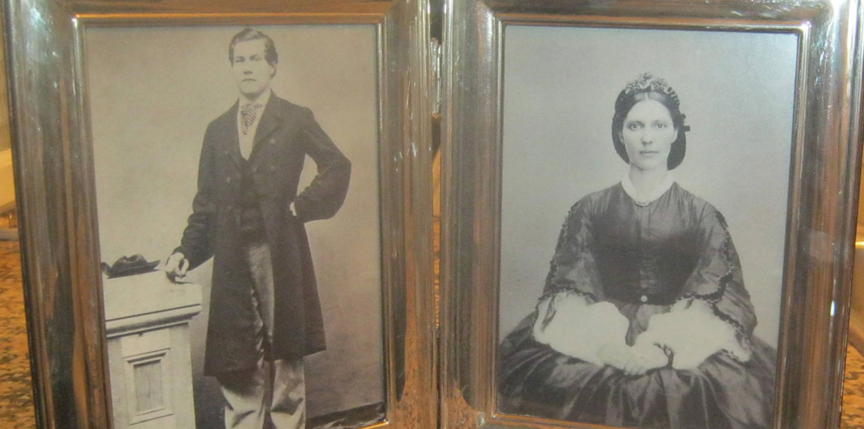 Black and white portrait of young woman with piercing brown eyes, wearing a dark dress with a broach and white sleeves, as well as head covering. Beside her is a second portrait beside her of a man with dark hair standing, wearing a dark suit, with his left arm poised on his hip.
