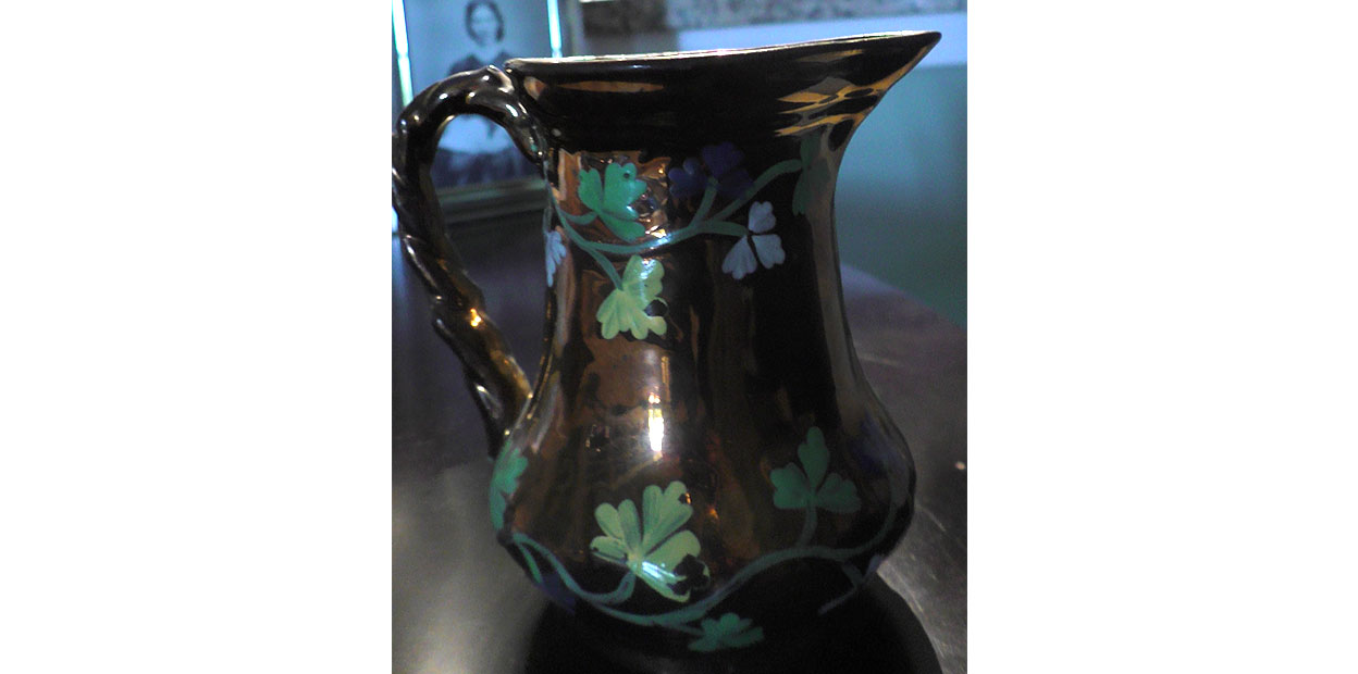 Close up photograph of gold coloured creamer with green shamrocks, with portrait of Bridget Ann Treay in background left.