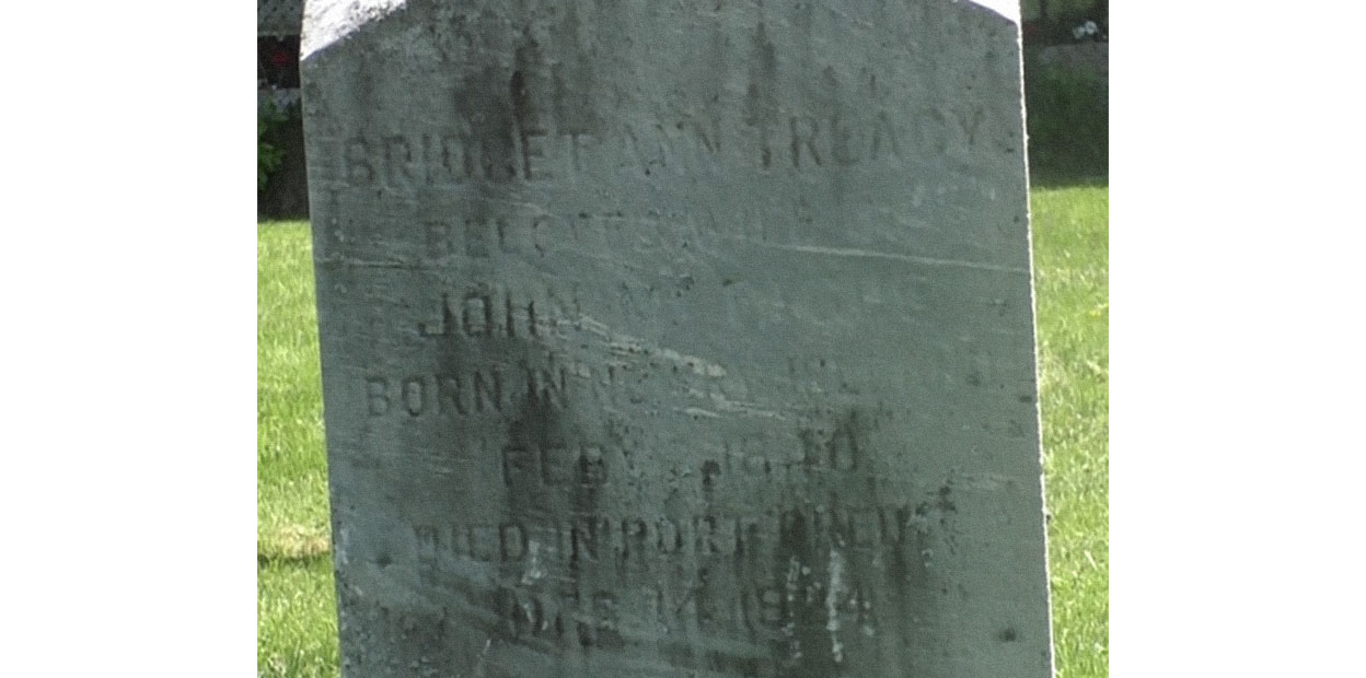 Close up photograph of grey grave marker with Bridget Ann Treacy visible on top.  Green grass and red brick building and windows visible in background.