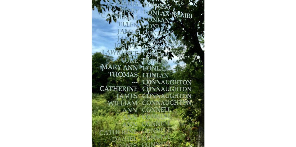 Texte inscrit en blanc, sur une surface vitrée qui reflète le ciel bleu et les branches d’un arbre. Le nom de Mary Conlan (Blair) est visible parmi ceux qui y sont inscrits.