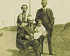Une photographie en noir et blanc où figurent quatre personnes, incluant un homme âgé et chauve, portant la moustache et vêtu d’un complet foncé, une jeune enfant habillée en blanc assise à l’avant, une femme aux courts cheveux foncés vêtue d’une robe et un homme portant la moustache et vêtu d’un complet, debout derrière eux. Un cheval est aperçu à l’arrière-plan, à droite.