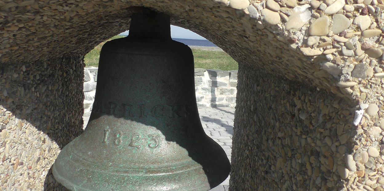 Grey bell mounted in monument with Carricks 1823 inscribed on it. Grey brick floor and wall, grass, and the sea visible in background.