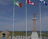 Wide angle view of two monuments, one with the bell featured earlier, and to its right a larger monument with a brown cross on grey plinth with three layers. Behind them an Irish, Canadian, and Quebec flag are flying in a moderate breeze, with a grey stone wall and blue sky visible in the background.