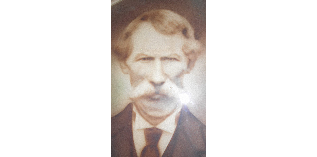 Une photographie en noir et blanc d’un homme portant une moustache blanche, de courts cheveux, bouclés près des oreilles, fixant la caméra. Il est vêtu d’un habit-cravate.