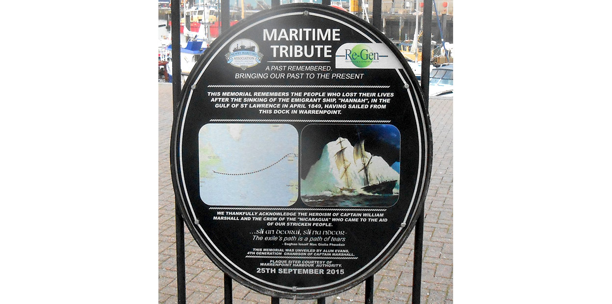 Circular plaque in black with white text and image of the Hannah striking an ice ber as well as map of its route from Ireland to Canada. Maritime Tribute is the heading on the plaque in larger text than interpretive script below. The plaque is mounted on a black railed fence with harbour and ship visible behind it.