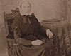 Black and white sepia toned photograph of seated woman in dark dress wearing white shawl staring into camera. To her right is a table covered with a cloth with a binder on it.
