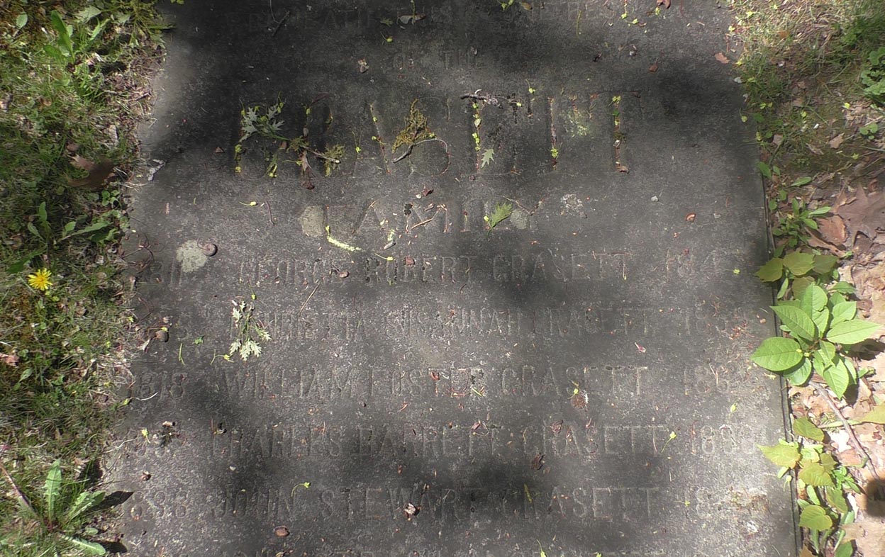 Photographie en plan rapproché d’une pierre tombale horizontale. Quelques feuilles et branches sont tombées sur l’épitaphe. Le nom de six personnes figurent sur la pierre tombale.
