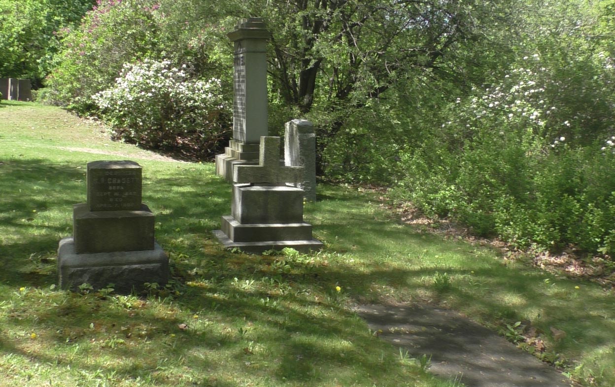 Horizontal grave slab, bottom right, with four vertical grave markers behind it, and row of trees and bushes marking cemetery boundary in background.