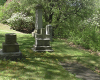 Pierre tombale horizontale aperçue dans le coin inférieur droit, ainsi que quatre autres monuments funéraires verticaux visibles plus loin derrière. Une rangée d’arbres et de buissons, marquant la limite du cimetière, sont aperçus à l’arrière-plan.