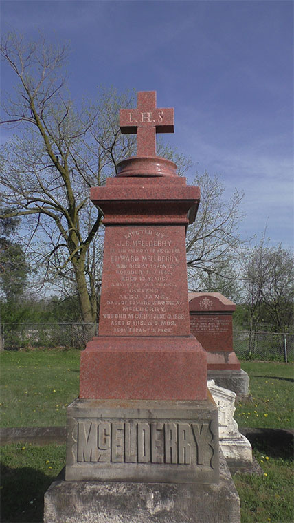 Red granite memorial marker with cross on top, mounted on grey stone plinth with McElderry inscribed on its base. Tree and grass in background.