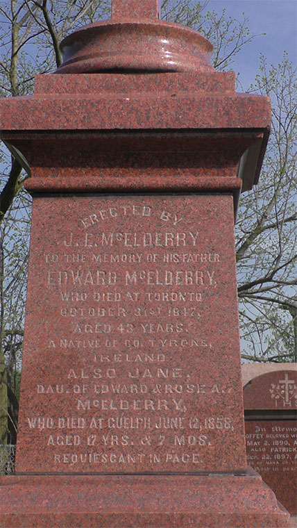 Plan rapproché de l’inscription lisible sur le monument de granite rouge. Des branches de l’arbre sont aperçues à l’arrière-plan.