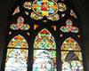 Stained glass windows, in shape of a circular flower with six petals and two shamrocks, at top, below which are three arched shaped windows with six figures depicted in them. Three rectangular windows below them with McElderry inscribed in centre one.