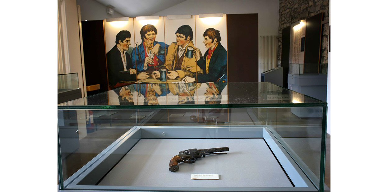 Photograph of a black pistol with a brown handle in a glass display case. Behind it can be seen an illuminated poster with four illustrated figures of men, wearing black, blue, yellow, and dark blue jackets. They are engaged in conversation, and one of them is lifting a mug.
