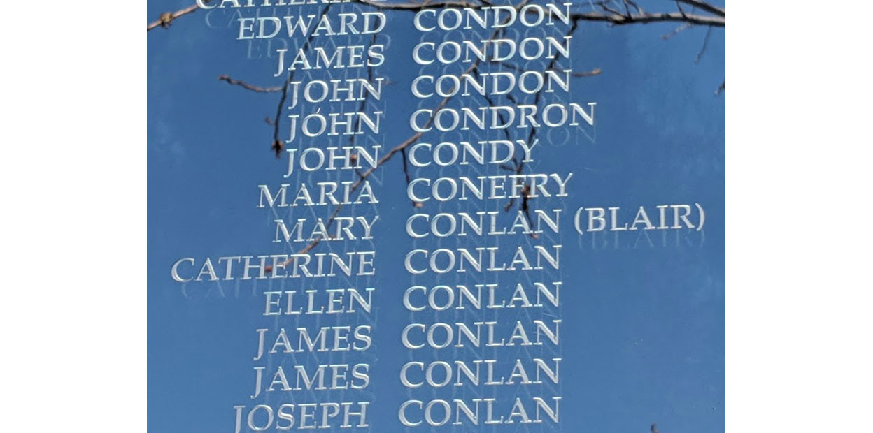 Texte inscrit en blanc, sur une surface vitrée qui reflète le ciel bleu et les branches d’un arbre. Le nom de Mary Conlan (Blair) est visible parmi ceux qui y sont inscrits.