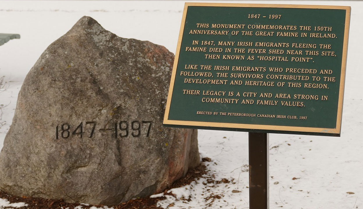 Large grey rock with 1847 – 1997 inscribed on it. A mounted plaque to its right has gold text on a green background that reads: 1847 – 1997. THIS MONUMENT COMMEMORATES THE 150TH ANNIVERSARY OF THE GREAT FAMINE IN IRELAND. IN 1847, MANY IRISH EMIGRANTS FLEEING THE FAMINE DIED IN THE FEVER SHED NEAR THIS SITE, THEN KNOWN AS HOSPITAL POINT. LIKE THE IRISH EMIGRANTS WHO PRECEDED AND FOLLOWED, THE SURVIVORS CONTRIBUTED TO THE DEVELOPMENT AND HERITAGE OF THIS REGION. THEIR LEGACY IS A CITY AND AREA STRONG IN COMMUNITY AND FAMILY VALUES. ERECTED BY THE PETERBOROUGH CANADIAN IRISH CLUB, 1997. Snow and frozen lake visible in background.