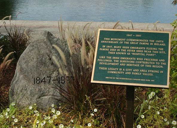 Large grey rock in foreground set in flowers and green grasse with 1847 – 1997 inscribed on, it beside a mounted plaque to its right that has gold text on a green background. Behind them can be seen body of water.