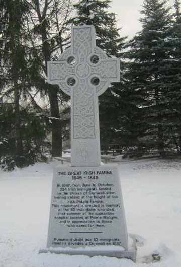 Grey Celtic Cross in snowy ground with several coniferous trees and river in background. Inscribed on its base: The Great Irish Famine. 1845-1848, with additional smaller inscriptions underneath.